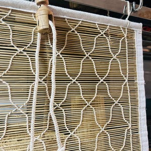 May include: A natural bamboo window shade with a woven pattern and a white fabric border. The shade features a wooden pulley system and a thick, off-white rope for raising and lowering. The bamboo slats are a light brown color.