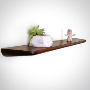 Puede incluir: Estante flotante de madera marrón oscuro con borde redondeado. Una maceta geométrica blanca y morada con una suculenta y una pequeña figura rosa se exhiben en el estante. El estante está montado en una pared blanca.