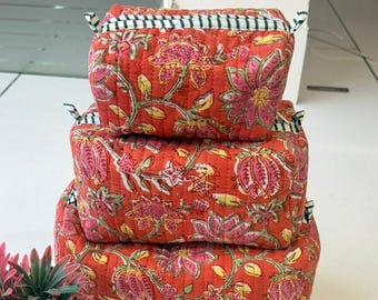 Puede incluir: Una pila de tres estuches rectangulares de tela acolchada de diferentes tamaños, con un fondo naranja vibrante y un patrón floral repetitivo en rosa y verde. Cada estuche tiene un cierre de cremallera con detalles de tela a rayas blancas y negras.