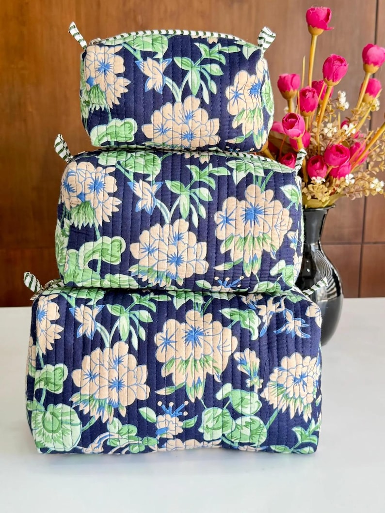 Puede incluir: Una pila de tres estuches acolchados rectangulares de diferentes tama&ntilde;os, hechos de tela azul marino oscuro con un patr&oacute;n floral repetitivo en tonos melocot&oacute;n claro, verde y azul. Cada estuche tiene una cremallera y una leng&uuml;eta de tela a rayas.