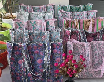 May include: A collection of quilted tote bags featuring various floral and striped patterns in shades of pink, blue, green, and purple. Each bag has two shoulder straps and a rectangular shape with visible stitching throughout the fabric surface.