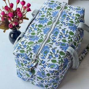 May include: A rectangular quilted duffel bag featuring a white base with a repeating blue and green floral pattern. The bag has a central zipper closure and black and white striped fabric handles attached to the sides for carrying.
