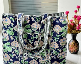 May include: A rectangular quilted tote bag featuring a dark navy blue background with a repeating floral pattern in shades of light pink, blue, and green. The bag has two matching fabric handles and is finished with a black and white striped trim.