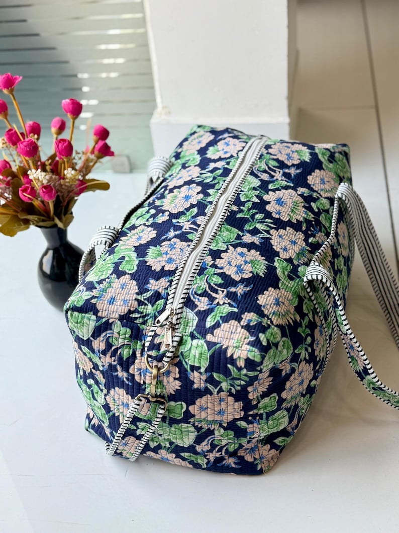 May include: A quilted duffel bag featuring a dark navy blue background with a repeating floral pattern in shades of light pink, green, and white. The bag includes a white zipper closure and two black and white striped fabric shoulder straps.
