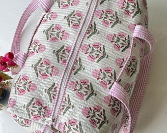 May include: A quilted duffel bag featuring a white base with a repeating pink and green floral block print pattern. The bag includes a white zip closure and two light pink and white striped fabric handles attached to the sides.