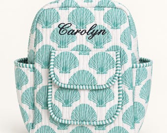 May include: A quilted backpack featuring a white background with a repeating teal seashell pattern. The bag includes a top handle, two side pockets, and a front flap pocket. The name Carolyn is embroidered in black script on the front panel.