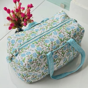May include: A quilted duffel bag featuring a white base with a repeating pattern of blue floral clusters and green leaves. The bag includes a top zipper closure and two light blue and white striped fabric handles attached to the sides.
