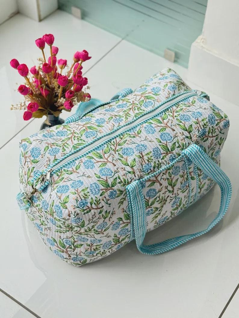 May include: A quilted duffel bag featuring a white base with a repeating pattern of blue floral clusters and green leaves. The bag includes a top zipper closure and two light blue and white striped fabric handles attached to the sides.