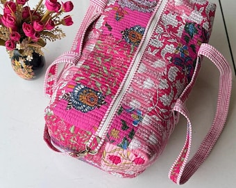 May include: A quilted duffel bag in vibrant shades of pink, featuring a detailed floral and botanical print throughout. The bag includes a white zip closure running along the top and two striped fabric handles for carrying.
