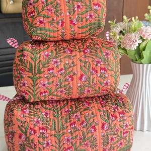 Puede incluir: Una pila de tres estuches rectangulares acolchados de tela color naranja melocotón con un patrón repetitivo de tallos verdes y flores rosas. Cada estuche tiene pequeñas lengüetas de tela a rayas rosas y blancas en las esquinas superiores.