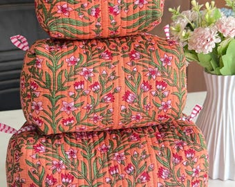 Puede incluir: Una pila de tres estuches rectangulares acolchados de tela color naranja melocotón con un patrón repetitivo de tallos verdes y flores rosas. Cada estuche tiene pequeñas lengüetas de tela a rayas rosas y blancas en las esquinas superiores.
