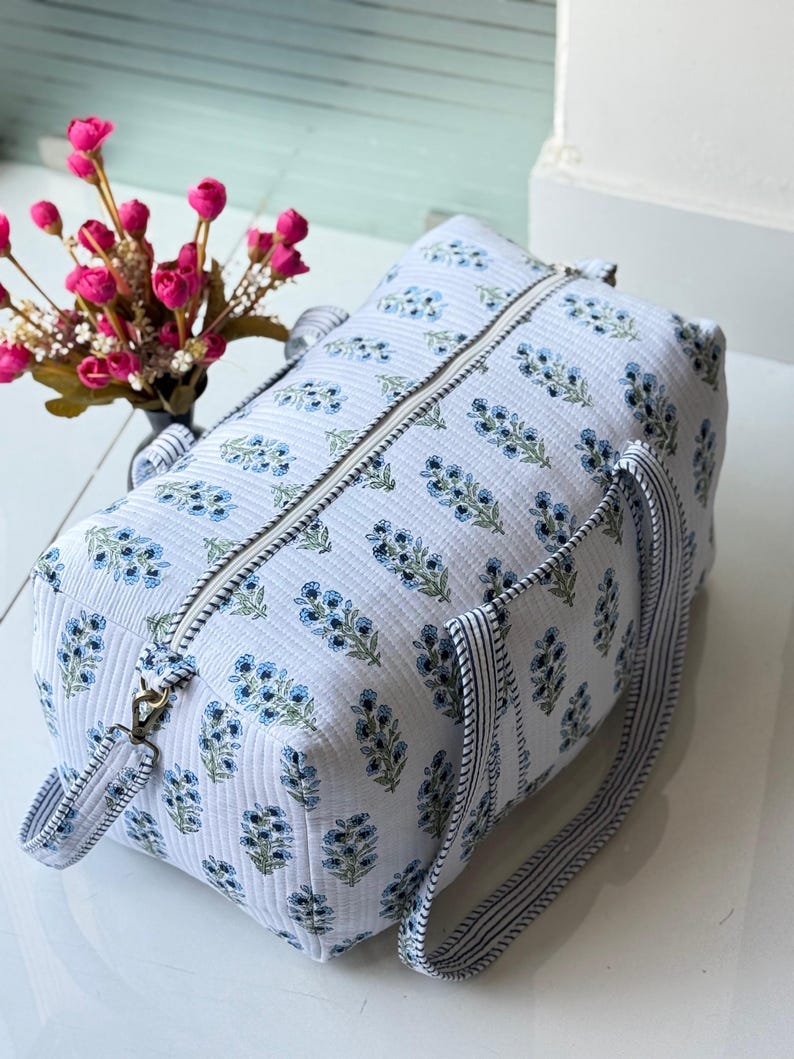 May include: A quilted duffel bag in white fabric featuring a repeating blue floral print. The bag has a top zipper closure, two striped carrying handles, and a detachable side strap with metal hardware. The texture is ribbed and soft-sided.