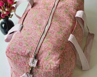 May include: A quilted duffel bag featuring a pink floral pattern with green leaves, a white zip closure, and light pink and white striped fabric handles. The bag has a rectangular shape with rounded edges and visible stitching throughout the fabric.