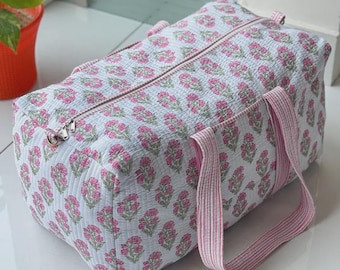 May include: A rectangular quilted duffel bag featuring a white base with a repeating pink floral block print pattern. The bag includes a top zip closure and two pink and white striped fabric handles attached to the sides.