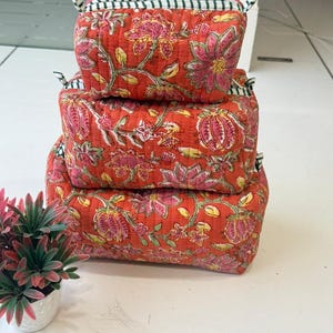 Puede incluir: Una pila de tres estuches rectangulares de tela acolchada de diferentes tama&ntilde;os, con un fondo naranja vibrante y un patr&oacute;n floral repetitivo en rosa y verde. Cada estuche tiene un cierre de cremallera con detalles de tela a rayas blancas y negras.