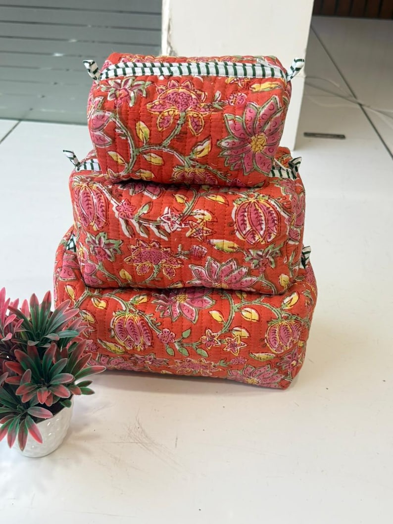 Puede incluir: Una pila de tres estuches rectangulares de tela acolchada de diferentes tama&ntilde;os, con un fondo naranja vibrante y un patr&oacute;n floral repetitivo en rosa y verde. Cada estuche tiene un cierre de cremallera con detalles de tela a rayas blancas y negras.