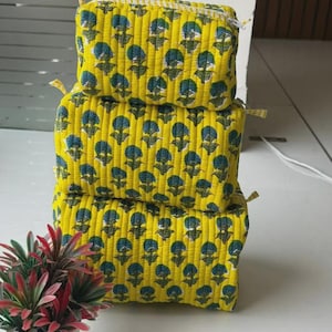 Puede incluir: Una pila de tres estuches rectangulares de tela acolchada en color amarillo brillante. Cada estuche presenta un patr&oacute;n repetitivo de motivos florales azul oscuro con tallos y hojas verdes. Los estuches tienen cierre de cremallera y costuras verticales.