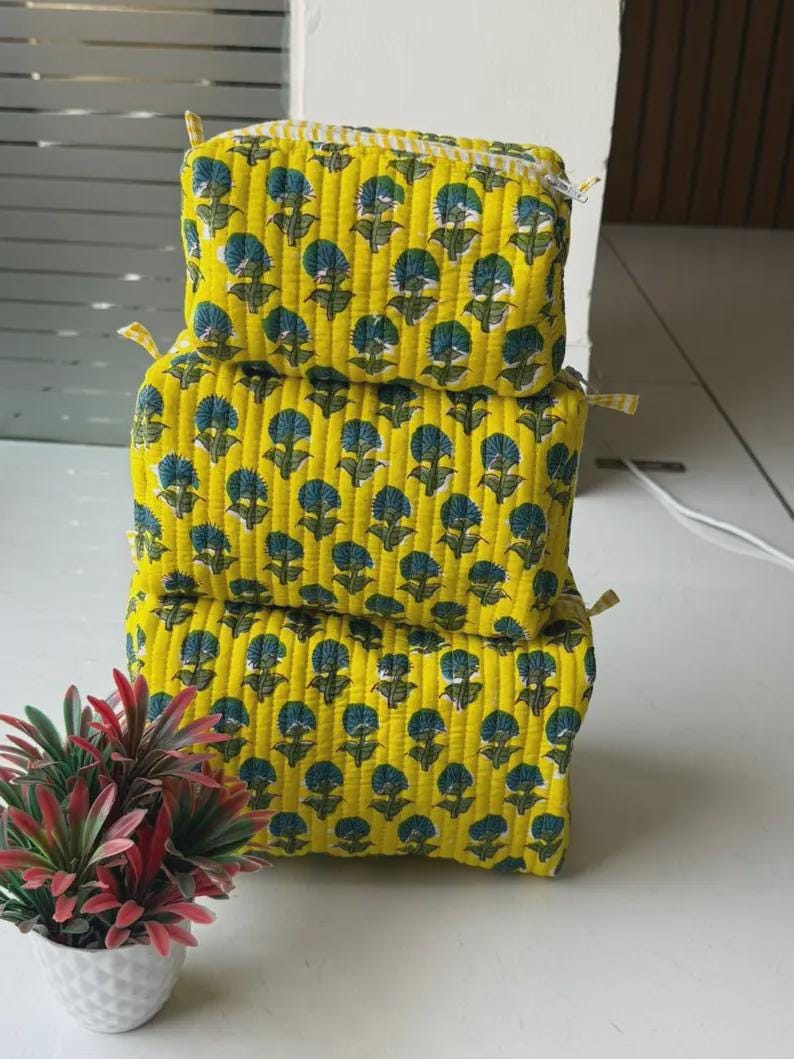 Puede incluir: Una pila de tres estuches rectangulares de tela acolchada en color amarillo brillante. Cada estuche presenta un patr&oacute;n repetitivo de motivos florales azul oscuro con tallos y hojas verdes. Los estuches tienen cierre de cremallera y costuras verticales.