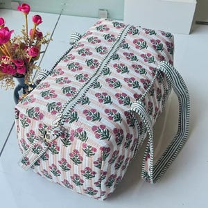 May include: A rectangular quilted duffel bag featuring a repeating pattern of small pink floral sprigs on a background of thin vertical peach and white stripes. The bag includes a top zipper closure and two striped fabric handles for carrying.