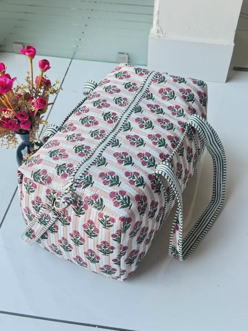 May include: A rectangular quilted duffel bag featuring a repeating pattern of small pink floral sprigs on a background of thin vertical peach and white stripes. The bag includes a top zipper closure and two striped fabric handles for carrying.