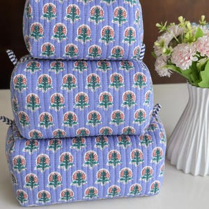 Puede incluir: Un conjunto de tres bolsas de almacenamiento rectangulares acolchadas apiladas verticalmente. Cada bolsa presenta una tela de color morado claro con un patr&oacute;n floral repetitivo en rosa y verde, acentuado por bordes de tela a rayas azules y blancas.