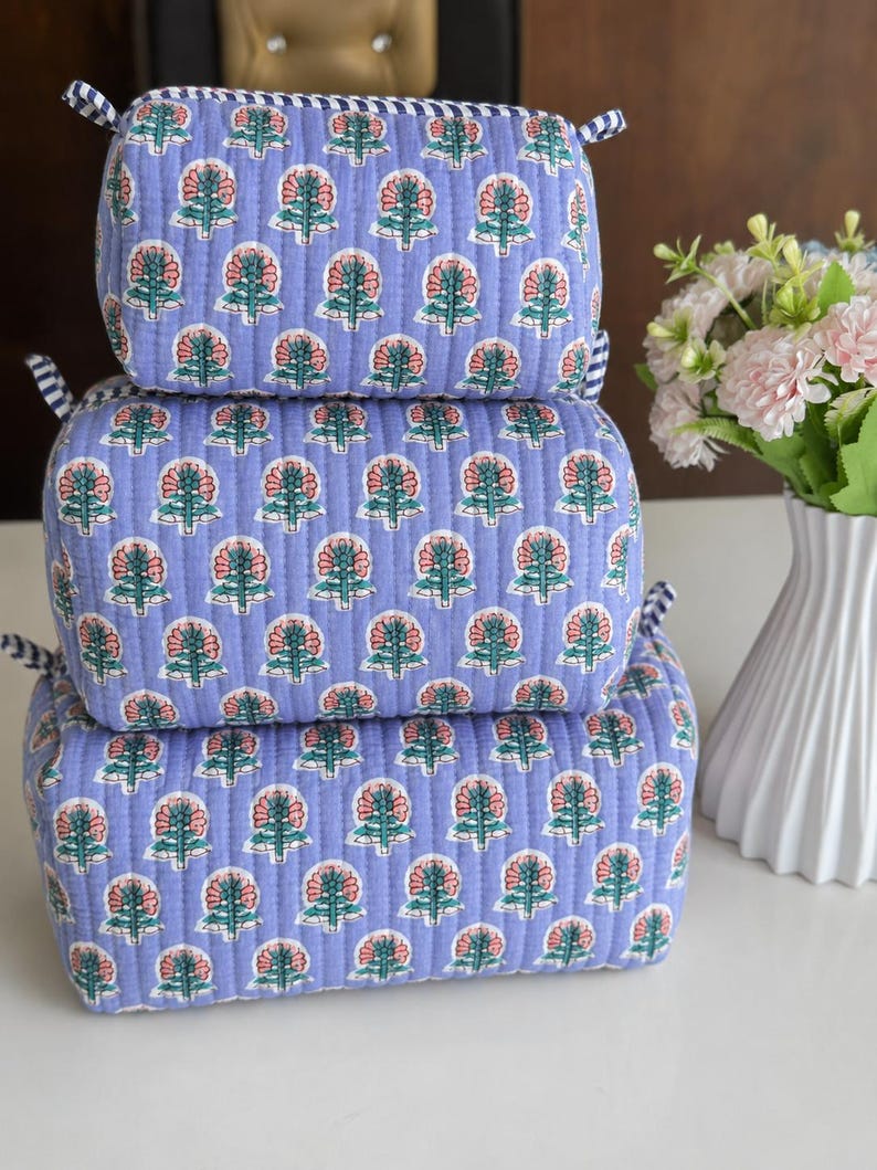 Puede incluir: Un conjunto de tres bolsas de almacenamiento rectangulares acolchadas apiladas verticalmente. Cada bolsa presenta una tela de color morado claro con un patr&oacute;n floral repetitivo en rosa y verde, acentuado por bordes de tela a rayas azules y blancas.
