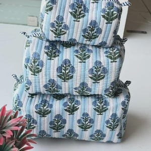 Può includere: Un set di tre pochette trapuntate rettangolari impilate verticalmente. Ogni pochette presenta un motivo a righe verticali azzurre e bianche con un motivo floreale ripetuto in blu e verde scuro. Texture morbida e imbottita.