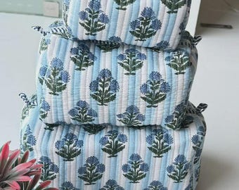 Puede incluir: Un conjunto de tres estuches acolchados rectangulares apilados verticalmente. Cada estuche presenta un patrón de rayas verticales en azul claro y blanco con un motivo floral repetitivo en azul y verde oscuro. Textura suave y acolchada.