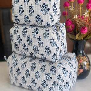 Puede incluir: Un conjunto de tres estuches acolchados rectangulares apilados verticalmente, cada uno con una base de tela blanca con un patr&oacute;n floral azul oscuro repetido, l&iacute;neas de costura verticales y un cierre de cremallera de metal plateado.