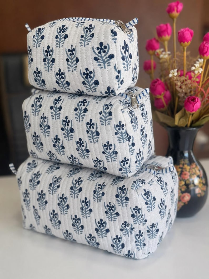 Puede incluir: Un conjunto de tres estuches acolchados rectangulares apilados verticalmente, cada uno con una base de tela blanca con un patr&oacute;n floral azul oscuro repetido, l&iacute;neas de costura verticales y un cierre de cremallera de metal plateado.