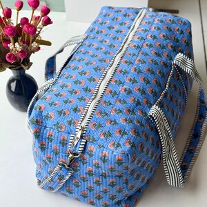 May include: A quilted duffel bag in a bright blue fabric featuring a repeating pattern of small pink flowers with green stems. The bag includes a white zipper closure along the top and two black and white striped fabric handles for carrying.