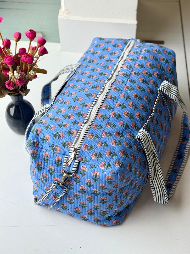 May include: A quilted duffel bag in a bright blue fabric featuring a repeating pattern of small pink flowers with green stems. The bag includes a white zipper closure along the top and two black and white striped fabric handles for carrying.