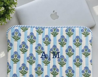 May include: A rectangular quilted laptop sleeve featuring a light blue and white vertical striped pattern with a repeating blue floral motif. The centre of the sleeve is embroidered with the dark blue capital letters A and M separated by a vertical line.