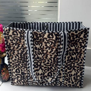 May include: A rectangular quilted tote bag featuring a brown and black leopard print pattern on the exterior. The bag has two black and white striped fabric handles and a matching black and white striped interior lining with a single snap button closure.