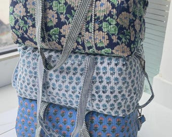 May include: A stack of three rectangular quilted duffel bags featuring floral patterns in navy blue, white, and light blue. Each bag has two striped fabric handles and a quilted texture throughout, showcasing distinct botanical prints on each piece.