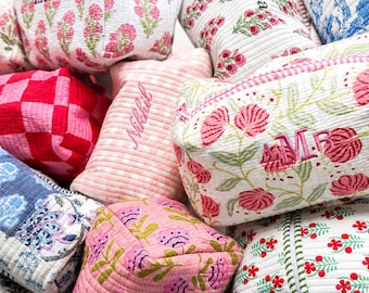 May include: A collection of quilted fabric pouches featuring various patterns including pink floral prints, red and pink checkered designs, and blue floral motifs. Several pouches are personalised with embroidered names such as Laura, Emma, Nikhil, and AMR.