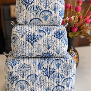 Puede incluir: Un conjunto de tres bolsas de almacenamiento rectangulares acolchadas apiladas verticalmente. Cada bolsa presenta una base de tela blanca con un patr&oacute;n azul repetitivo de conchas en forma de abanico y motivos de coral, con peque&ntilde;as pesta&ntilde;as a cuadros.