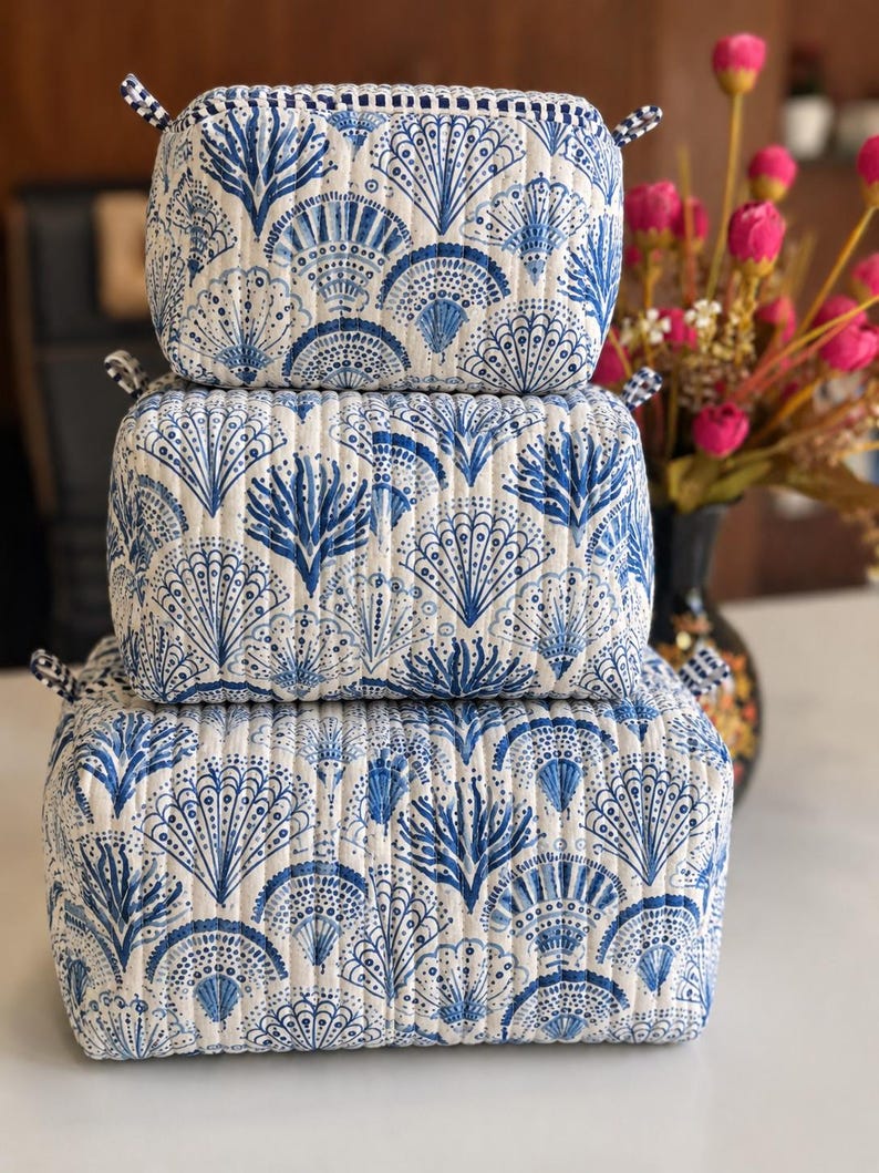 Puede incluir: Un conjunto de tres bolsas de almacenamiento rectangulares acolchadas apiladas verticalmente. Cada bolsa presenta una base de tela blanca con un patr&oacute;n azul repetitivo de conchas en forma de abanico y motivos de coral, con peque&ntilde;as pesta&ntilde;as a cuadros.