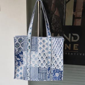 May include: A quilted tote bag featuring a patchwork design of various blue and white floral and striped patterns. The bag has two long shoulder straps and a rectangular shape, constructed with vertical stitching throughout the fabric surface.