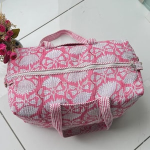 May include: A quilted duffel bag featuring a vibrant pink and white floral block print pattern. The bag includes two top handles with a pink and white striped design and a white zipper closure running across the top of the rectangular structure.