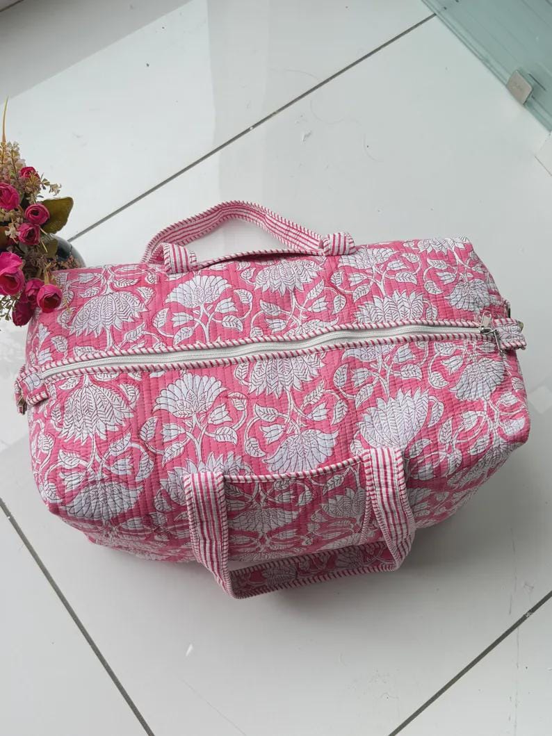 May include: A quilted duffel bag featuring a vibrant pink and white floral block print pattern. The bag includes two top handles with a pink and white striped design and a white zipper closure running across the top of the rectangular structure.
