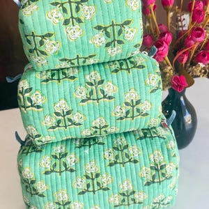 Puede incluir: Una pila de tres estuches rectangulares acolchados de tela verde claro con un patr&oacute;n floral repetitivo en blanco y amarillo sobre tallos verde oscuro. Cada estuche tiene una cremallera y un borde de tela a cuadros azules y blancos.