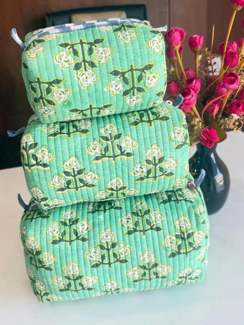 Puede incluir: Una pila de tres estuches rectangulares acolchados de tela verde claro con un patr&oacute;n floral repetitivo en blanco y amarillo sobre tallos verde oscuro. Cada estuche tiene una cremallera y un borde de tela a cuadros azules y blancos.
