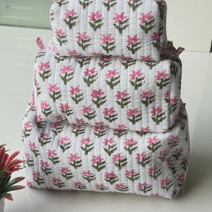 Puede incluir: Un conjunto de tres estuches acolchados rectangulares apilados verticalmente. Cada estuche es blanco con un patr&oacute;n repetitivo de flores rosas y tallos verdes. El estuche superior presenta un borde de cremallera a rayas rosas y blancas.