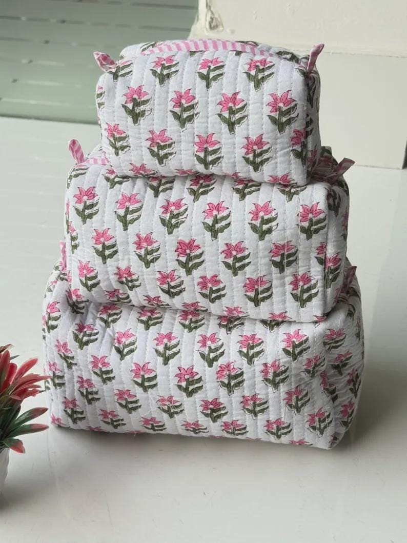 Puede incluir: Un conjunto de tres estuches acolchados rectangulares apilados verticalmente. Cada estuche es blanco con un patr&oacute;n repetitivo de flores rosas y tallos verdes. El estuche superior presenta un borde de cremallera a rayas rosas y blancas.