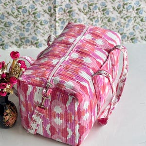 May include: A rectangular quilted duffel bag featuring a vibrant pink, white, and orange ikat-style pattern. The bag includes a top zipper closure, two side handles, and a detachable shoulder strap with metal hardware clips attached to the sides.
