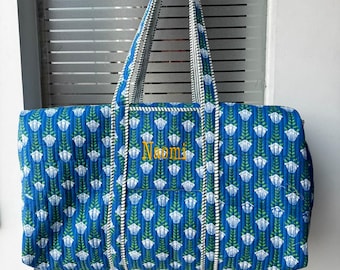 May include: A quilted duffel bag in a vibrant blue colour featuring a repeating pattern of white flowers with green stems. The bag has two long handles and a detachable shoulder strap with a black and white striped design. The name Naomi is embroidered in yellow.