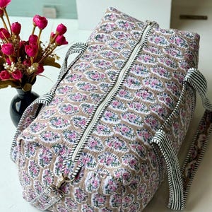 May include: A quilted duffel bag featuring a repeating floral pattern in shades of pink, blue, and green on a tan background. The bag has a white central zipper and black and white striped fabric handles attached with metal hardware.