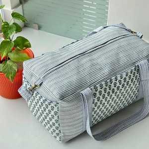 May include: A rectangular duffel bag featuring a blue and white striped pattern on the main body and handles, complemented by side panels adorned with a small green floral block print design, complete with a top zipper closure and metal hardware.
