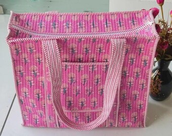 May include: A rectangular quilted tote bag in bright pink featuring a repeating pattern of small dark blue floral motifs. The bag has a zipped top closure, two matching pink and white striped fabric handles, and a front exterior pocket.